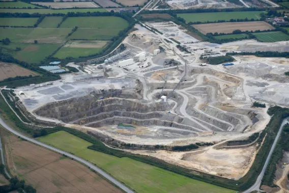 Carrière à ciel ouvert vue du ciel, avec des terrasses d’extraction, des routes sinueuses, des engins industriels et des zones de stockage de matériaux, entourée de champs agricoles