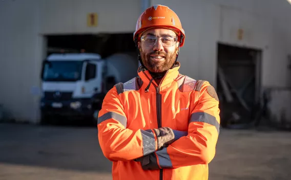 Collaborateur Nexstone en tenue de sécurité haute visibilité, bras croisés sur site industriel, avec véhicules et zone de production en arrière-plan.