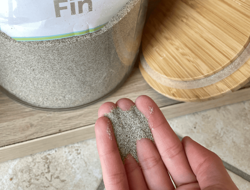 Sable fin 0/1 présenté dans une main, montrant sa granulométrie très fine et sa couleur gris clair, avec un contenant de produit en arrière-plan.