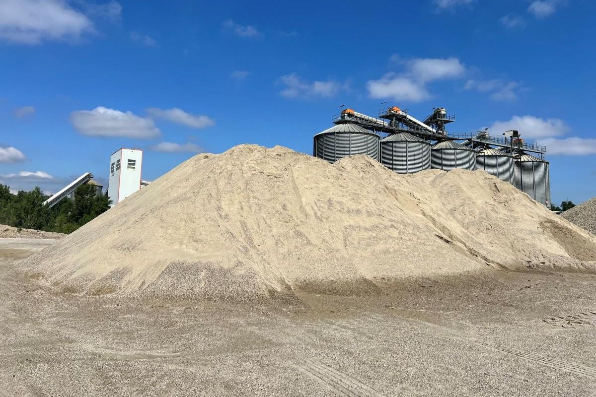 Stock important de sable clair entreposé dans la carrière Nexstone de Mions, avec des silos industriels en arrière-plan sous un ciel dégagé.