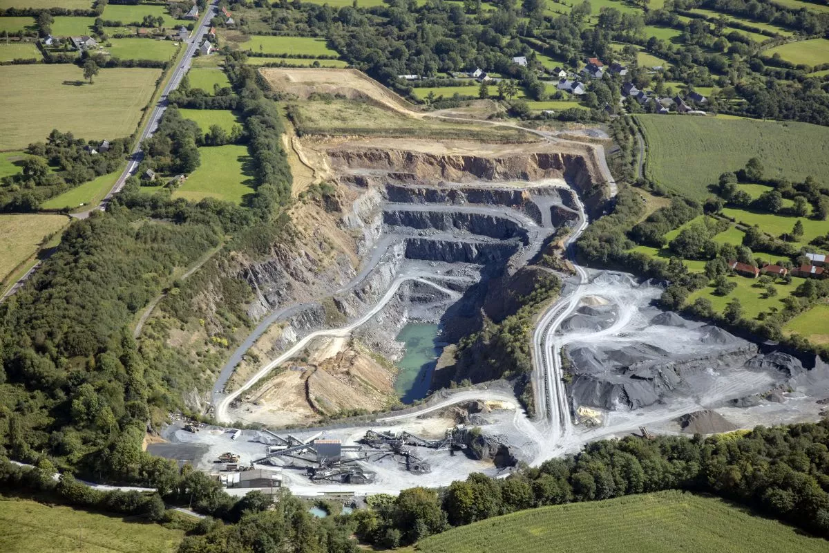 Vue aérienne de la carrière de Montsurvent, avec ses fronts de taille en gradins, ses installations de traitement et ses stocks de granulats, entourée de champs et de haies.