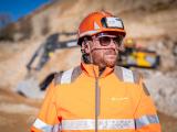 Conducteur d'engin sur la carrière de Brissac portant des équipements de protection (casque, lunettes, veste haute visibilité), sur site d’extraction de granulats avec engins de chantier en arrière-plan.