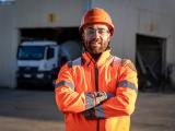 Collaborateur Nexstone en tenue de sécurité haute visibilité, bras croisés sur site industriel, avec véhicules et zone de production en arrière-plan.