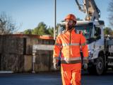 Opérateur en centrale à béton portant des équipements de protection (casque, tenue haute visibilité) se déplaçant sur site de production, avec installations de stockage et camion toupie en arrière-plan.